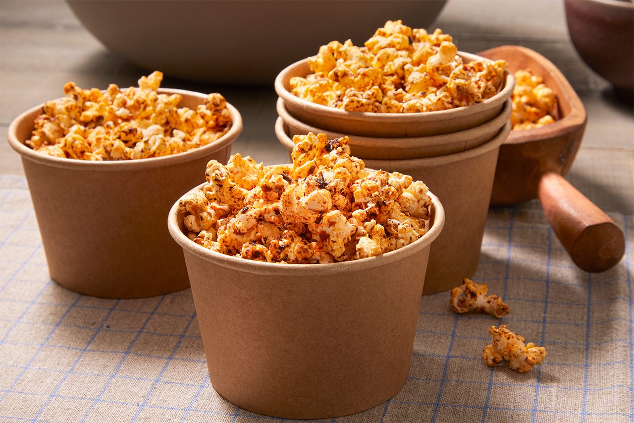 Three brown paper cups filled with seasoned popcorn are placed on a checkered surface, with a wooden scoop containing more popcorn in the background.
