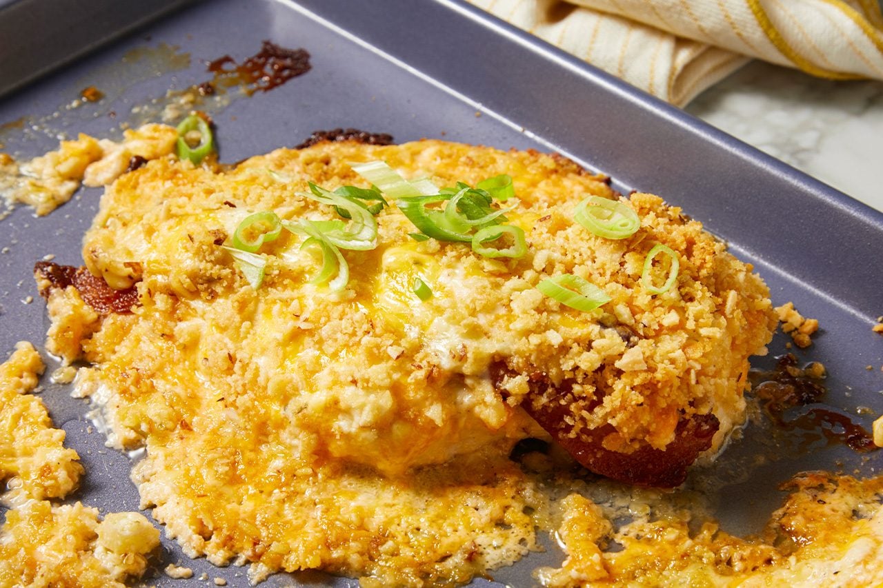 Eye Level horizontal image of a baked dish on a baking sheet. The dish, likely chicken, is covered with melted cheese and a crispy breadcrumb-like topping, garnished with sliced green onions. Melted cheese and some browned residue are visible on the baking sheet around the main item.