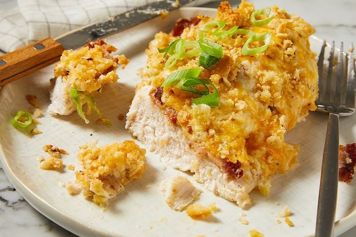 Eye Level horizontal image of a serving of oven-baked chicken, generously topped with a golden-brown, crispy crust, melted cheese, and bacon bits. The dish is garnished with fresh green onion slices and served on a light-colored plate with a knife and fork beside it.