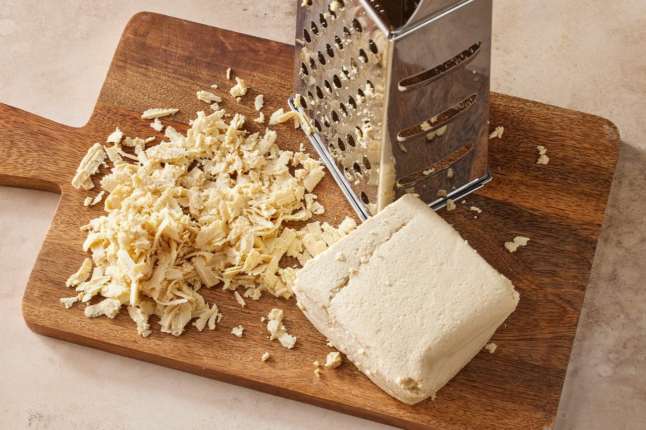 Overhead horizontal image of a block of firm tofu being grated on a metal box grater. A pile of shredded tofu rests on a dark wooden cutting board, alongside the grater and the remaining block of tofu. | [Original] Drain tofu on a kitchen towel for 10 minutes. Using the largest holes on a box grater, gently shred tofu. Push any large chunks that crumble off against the holes to shred.