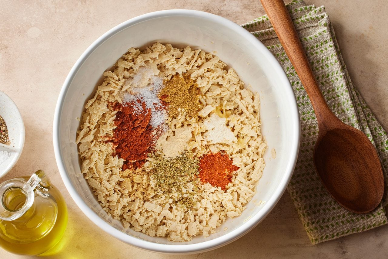 Overhead horizontal image of a white bowl containing a light-colored, crumbly mixture. Various spices, including salt, paprika, cumin, garlic powder, and dried oregano, are neatly piled on top, ready to be mixed. A cruet of olive oil, a small dish of salt and black pepper, and a wooden spoon resting on a green-dotted kitchen towel are visible on the sides. | [Original] Preheat oven to 400°. In a large bowl, combine shredded tofu, olive oil, soy sauce, chili powder, cumin, smoked paprika, garlic powder, onion powder, salt and oregano. Mix until tofu is thoroughly seasoned.