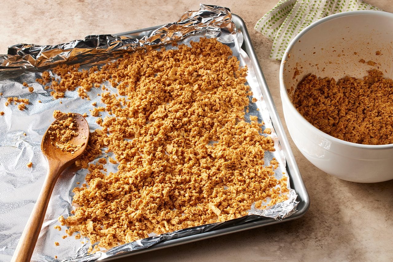 Overhead horizontal image of a crumbly golden-brown mixture spread out on an aluminum foil-lined baking sheet, with a wooden spoon resting on the foil. To the right, a white mixing bowl holds more of the same crumbly mixture, with a patterned kitchen towel visible behind it. | [Original] Spread tofu in a single layer on a foil-lined baking sheet sprayed with cooking oil; bake for 15 minutes.