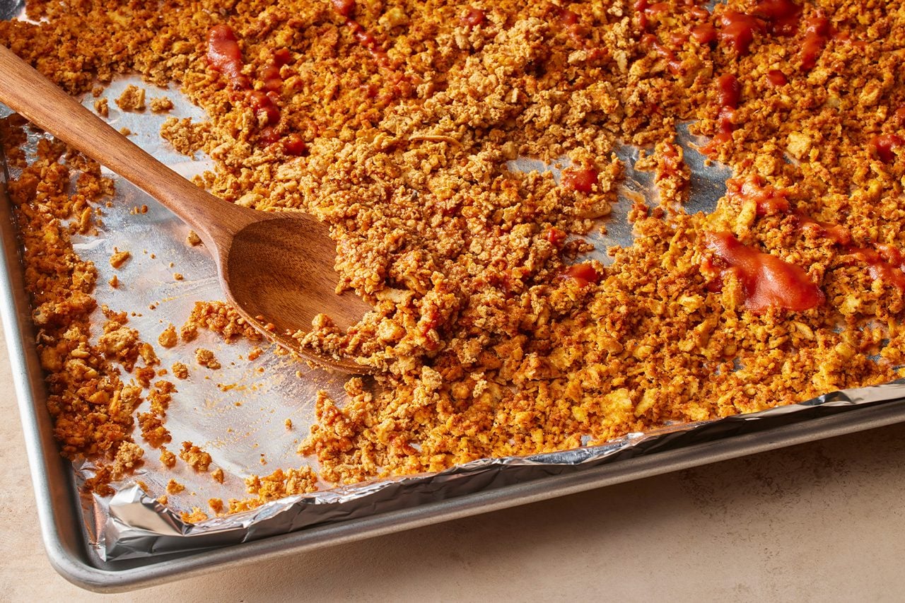 Overhead horizontal image of a baking sheet lined with aluminum foil, filled with a golden-brown crumbly mixture. A wooden spoon rests on the mixture, which has streaks of red sauce visible throughout. | [Original] In a small bowl, combine tomato paste with warm water. Remove the tofu from oven and drizzle with tomato water mixture; toss to coat.