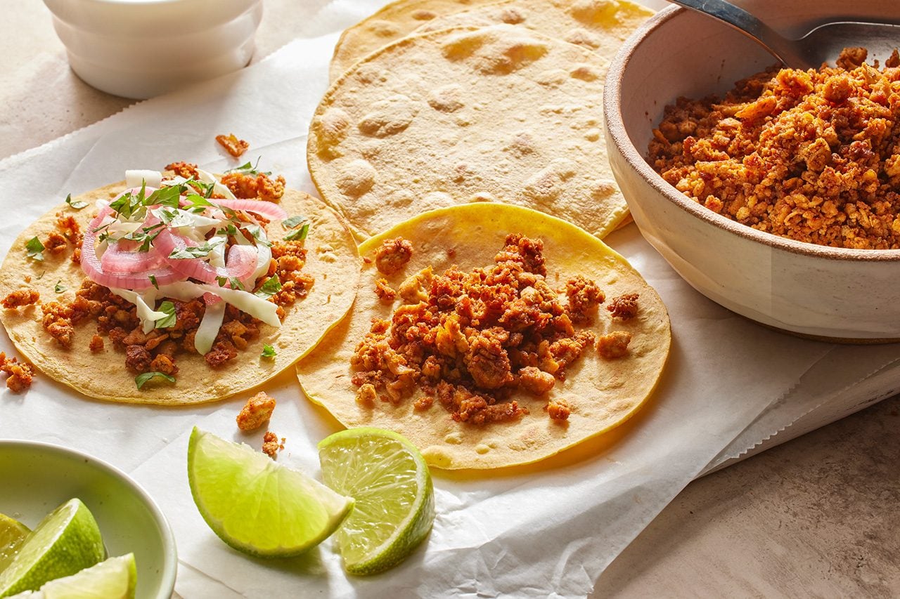 Eye level horizontal image of a spread of taco ingredients being assembled on white parchment paper. Two corn tortillas are filled with seasoned ground meat, one of which is topped with pickled red onions, shredded cabbage, and fresh cilantro. Lime wedges and a bowl of additional seasoned meat are also visible.