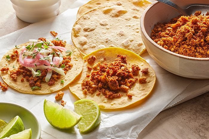 Eye level horizontal image of a spread of taco ingredients being assembled on white parchment paper. Two corn tortillas are filled with seasoned ground meat, one of which is topped with pickled red onions, shredded cabbage, and fresh cilantro. Lime wedges and a bowl of additional seasoned meat are also visible.
