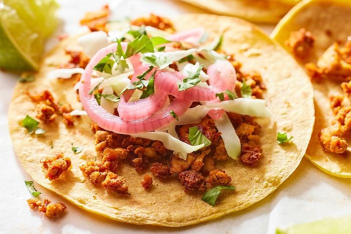 Eye-level horizontal image of a delicious taco. The taco features a corn tortilla filled with seasoned ground meat, topped with pickled red onions, shredded cabbage, and fresh green herbs; a lime wedge is visible to the left.