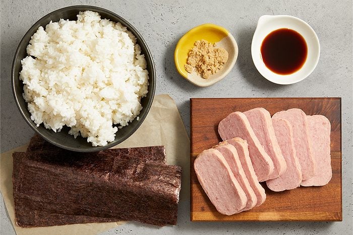 Overhead horizontal image of ingredients laid out on a gray countertop. A black bowl of white rice is positioned next to sheets of dark green nori on parchment paper. Slices of processed meat rest on a wooden cutting board, with small dishes of brown sugar and soy sauce nearby.