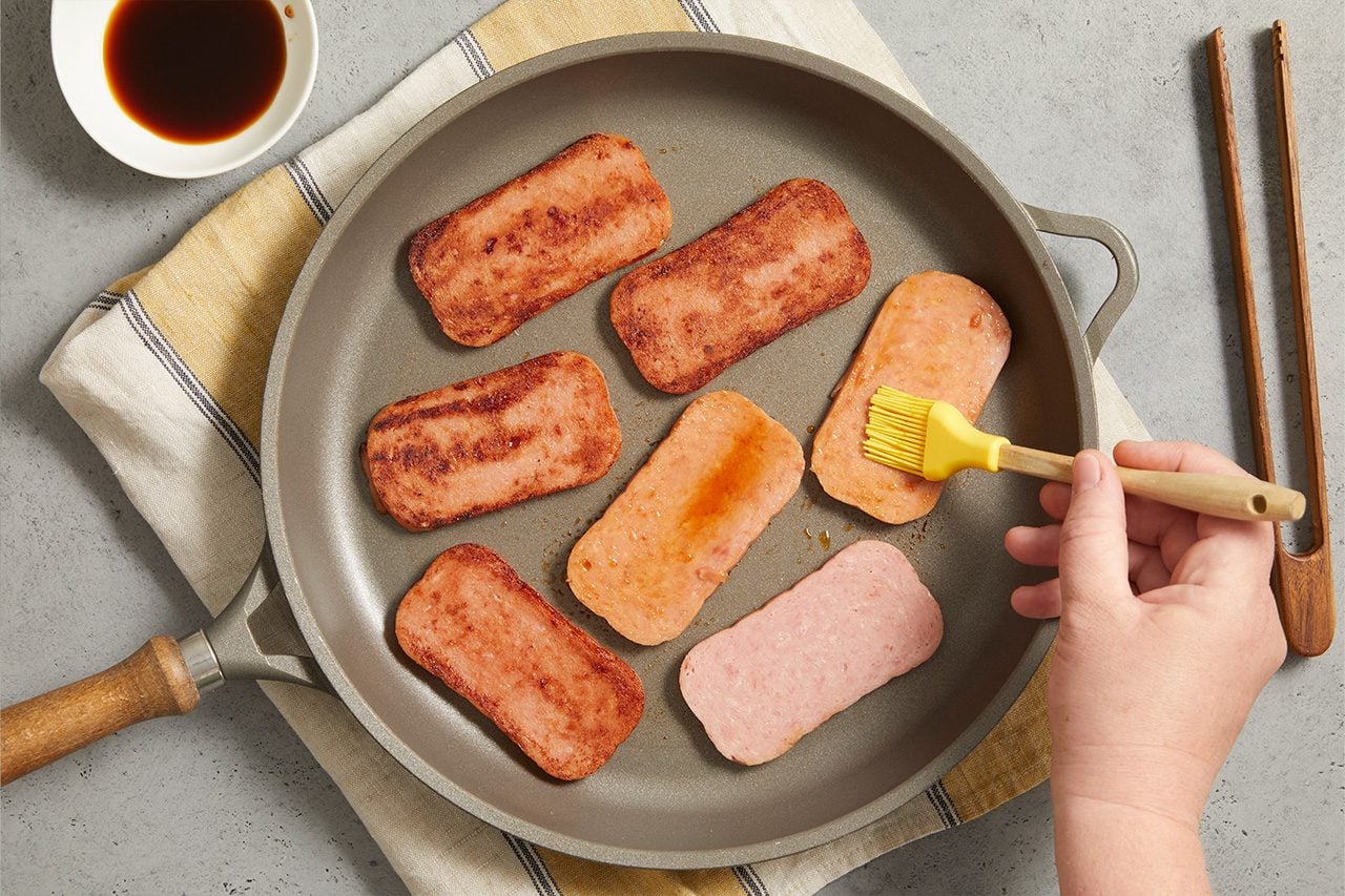 Overhead horizontal image of a hand brushing a glaze onto slices of Spam in a frying pan. Some of the processed meat slices are already browned and caramelized, while others are still pale. A small white bowl containing a dark sauce sits on a striped kitchen towel next to the pan.