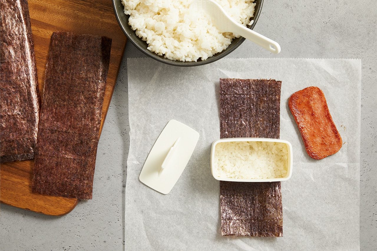 Overhead horizontal image of ingredients arranged on parchment paper, preparing to make a dish. A strip of nori seaweed is laid out with a rectangular mold filled with cooked white rice on top. Beside it, a slice of cooked spam rests, with a bowl of cooked rice and a paddle in the background.