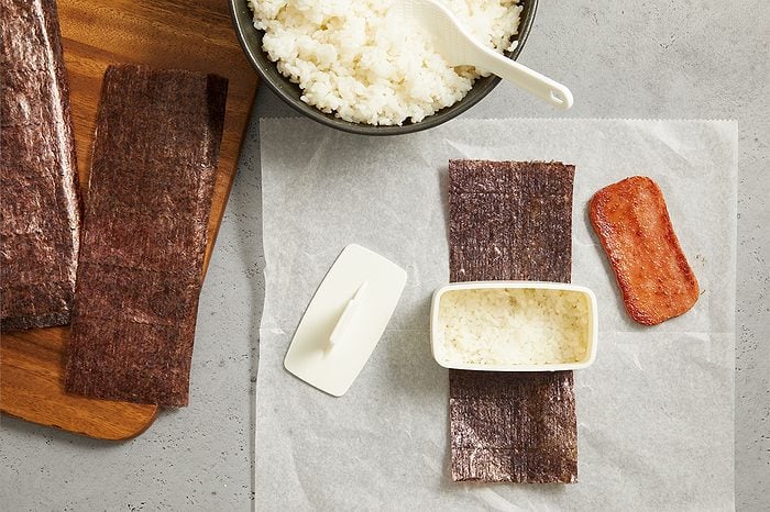 Overhead horizontal image of ingredients arranged on parchment paper, preparing to make a dish. A strip of nori seaweed is laid out with a rectangular mold filled with cooked white rice on top. Beside it, a slice of cooked spam rests, with a bowl of cooked rice and a paddle in the background.