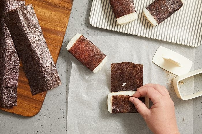 Overhead horizontal image of a hand wrapping a block of cooked rice with a nori sheet on parchment paper. Several other rice blocks wrapped in nori are visible, with some on a striped plate. Unused nori sheets rest on a wooden cutting board, alongside white plastic rice mold tools.