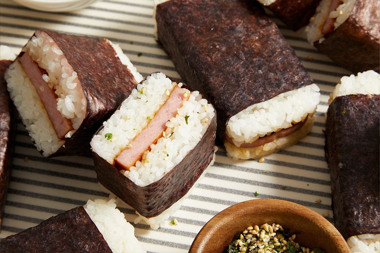 Overhead horizontal image of several freshly made Spam musubi arranged on a striped surface. The musubi are made with blocks of rice and grilled spam, wrapped in dark nori seaweed, with some cut open to show the layers. A small wooden bowl containing furikake, a Japanese seasoning blend of sesame seeds and dried seaweed, is visible in the foreground.