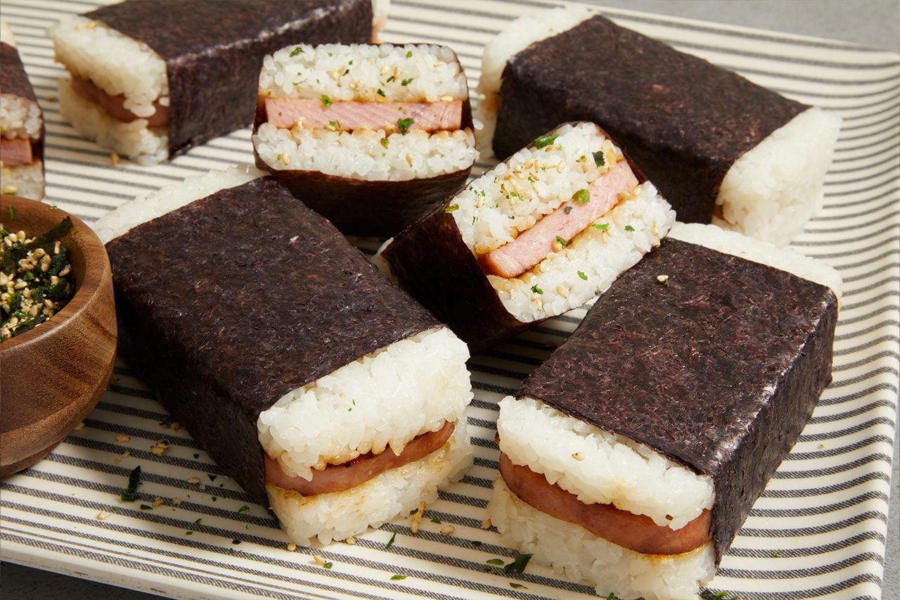 Eye Level horizontal image of several rectangular Spam musubi arranged on a striped platter. Some musubi are cut in half, revealing layers of white rice and sliced Spam. A small wooden bowl of furikake is visible on the left, with some sprinkled over the musubi and plate.