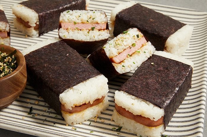Eye Level horizontal image of several rectangular Spam musubi arranged on a striped platter. Some musubi are cut in half, revealing layers of white rice and sliced Spam. A small wooden bowl of furikake is visible on the left, with some sprinkled over the musubi and plate.
