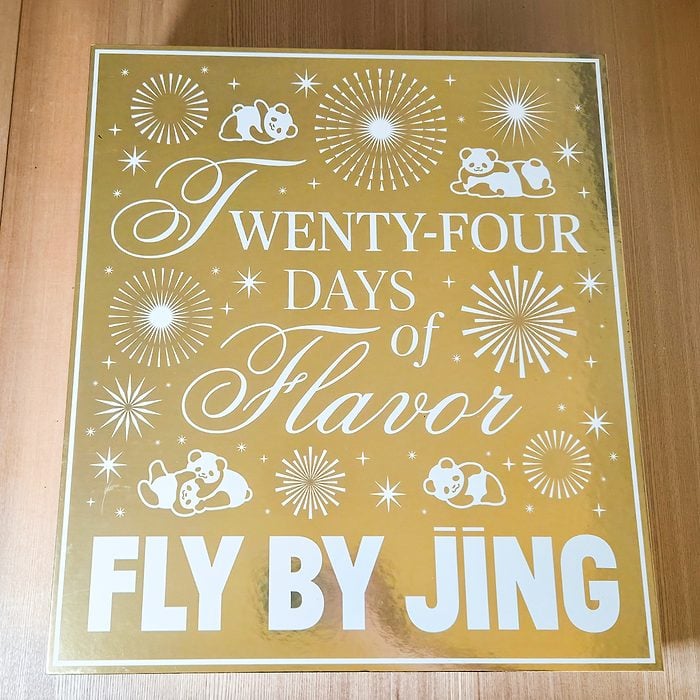 A gold box with white text that reads “TWENTY-FOUR DAYS of Flavor FLY BY JING,” decorated with fireworks and cartoon pandas.