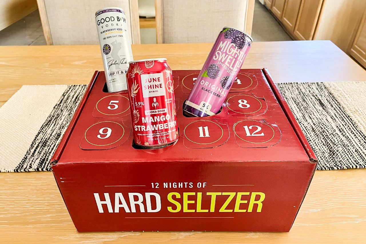 A red box labeled “12 Nights of Hard Seltzer” sits on a table, with three different hard seltzer cans placed on top of it, each with distinct designs and colors.