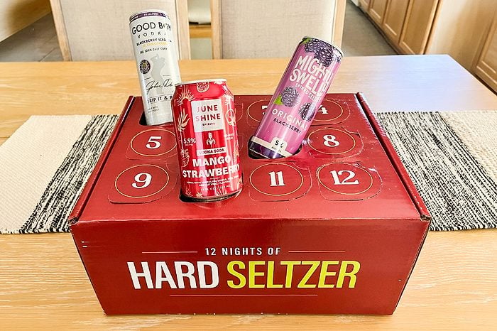 A red box labeled “12 Nights of Hard Seltzer” sits on a table, with three different hard seltzer cans placed on top of it, each with distinct designs and colors.
