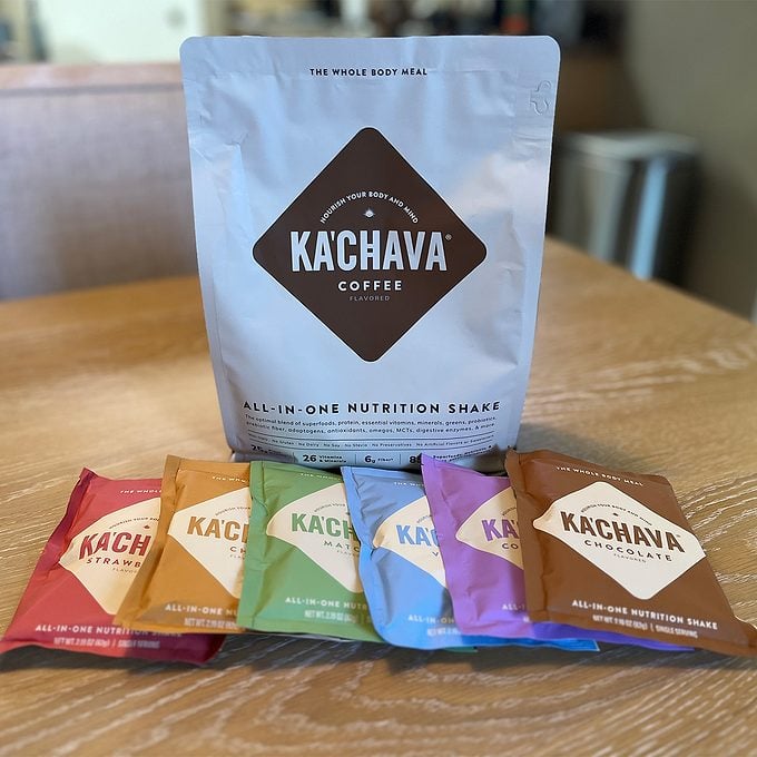 Several Ka’Chava all-in-one nutrition shake packets in different colors are arranged on a table, with a large Ka’Chava coffee-flavored bag standing behind them.