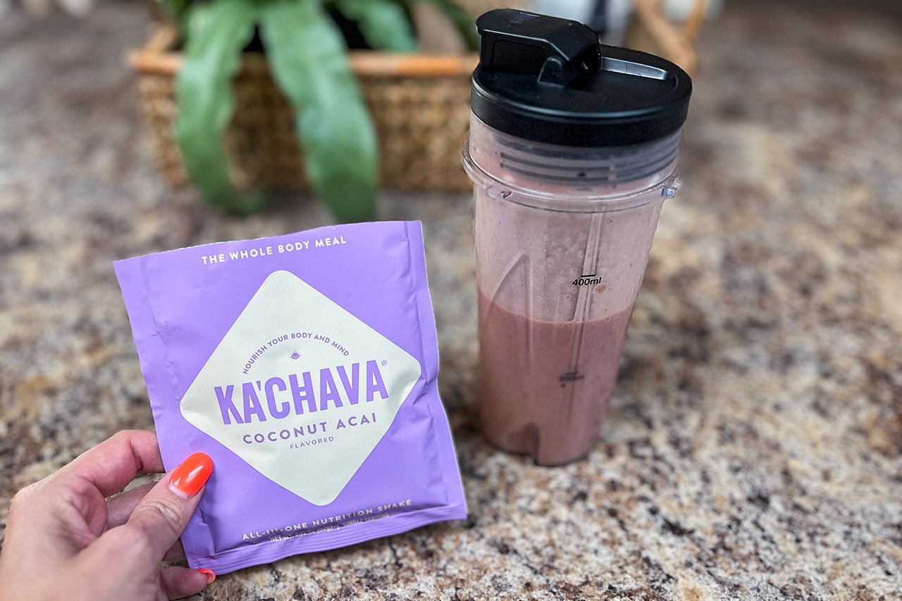 A hand holds a purple Ka’Chava Coconut Acai packet next to a blender bottle filled with a pink smoothie, on a brown speckled countertop.