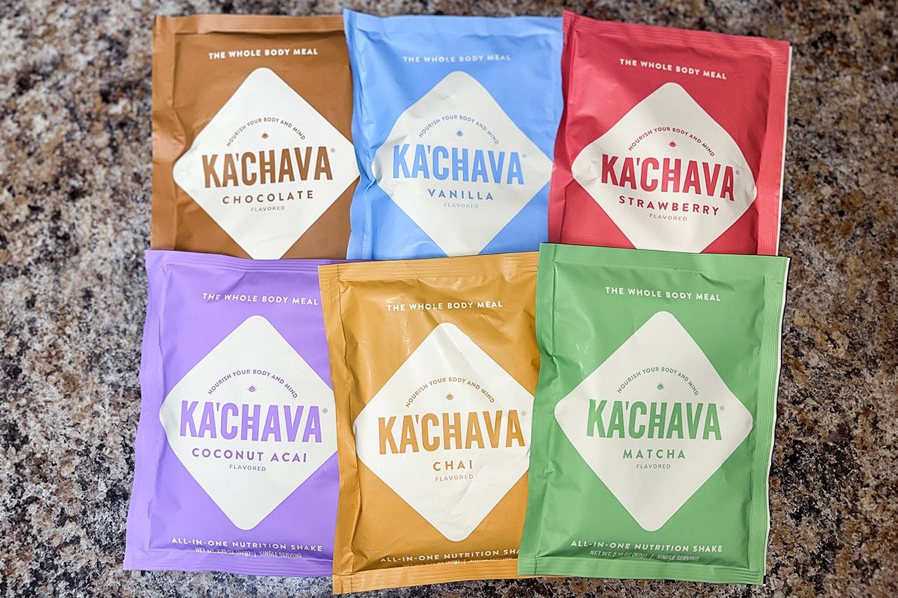 Six Kachava drink mix packets in different flavors—chocolate, vanilla, strawberry, coconut acai, chai, and matcha—arranged on a speckled countertop.