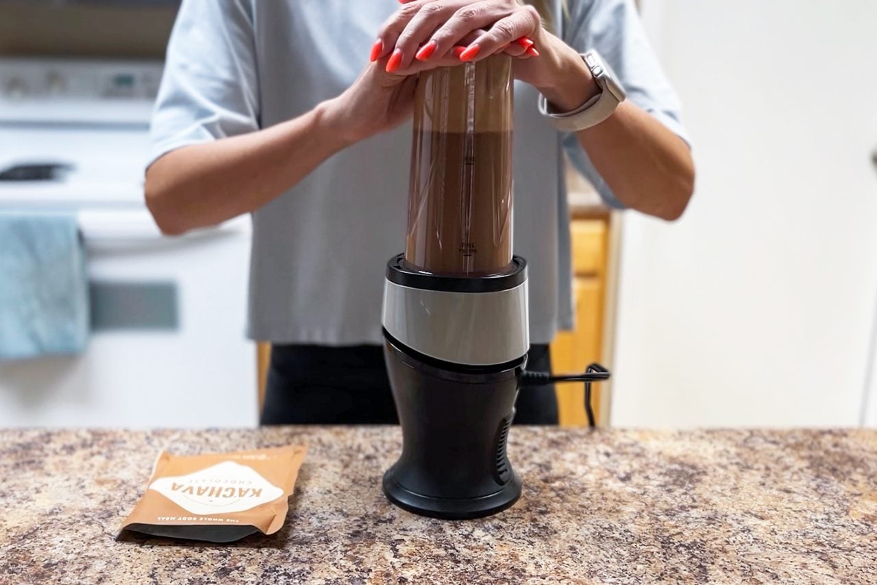 A person uses both hands to hold down a blender filled with a brown liquid on a kitchen counter. A brown pouch sits nearby. The background shows a stove and cabinets.
