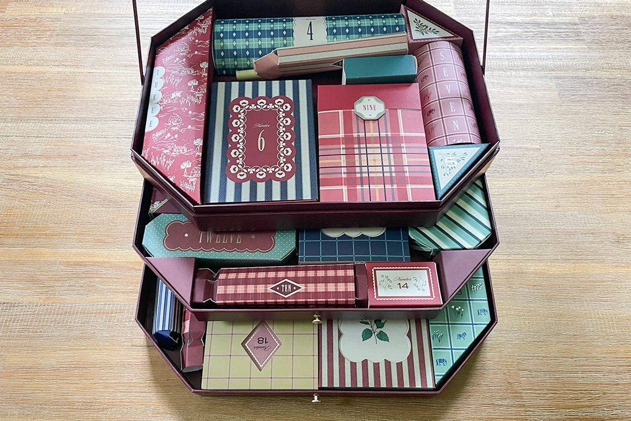 A two-tiered box holds various colorful, patterned rectangular packages, cards, and small gifts, neatly organized on each level against a wooden background.
