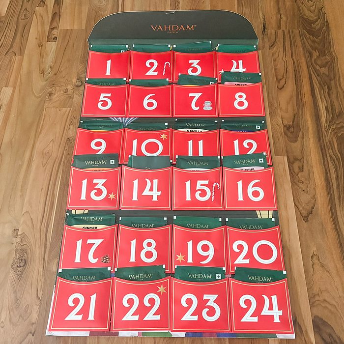 A Vahdam tea advent calendar with 24 red numbered doors is displayed on a wooden floor, each door representing a day leading up to Christmas.