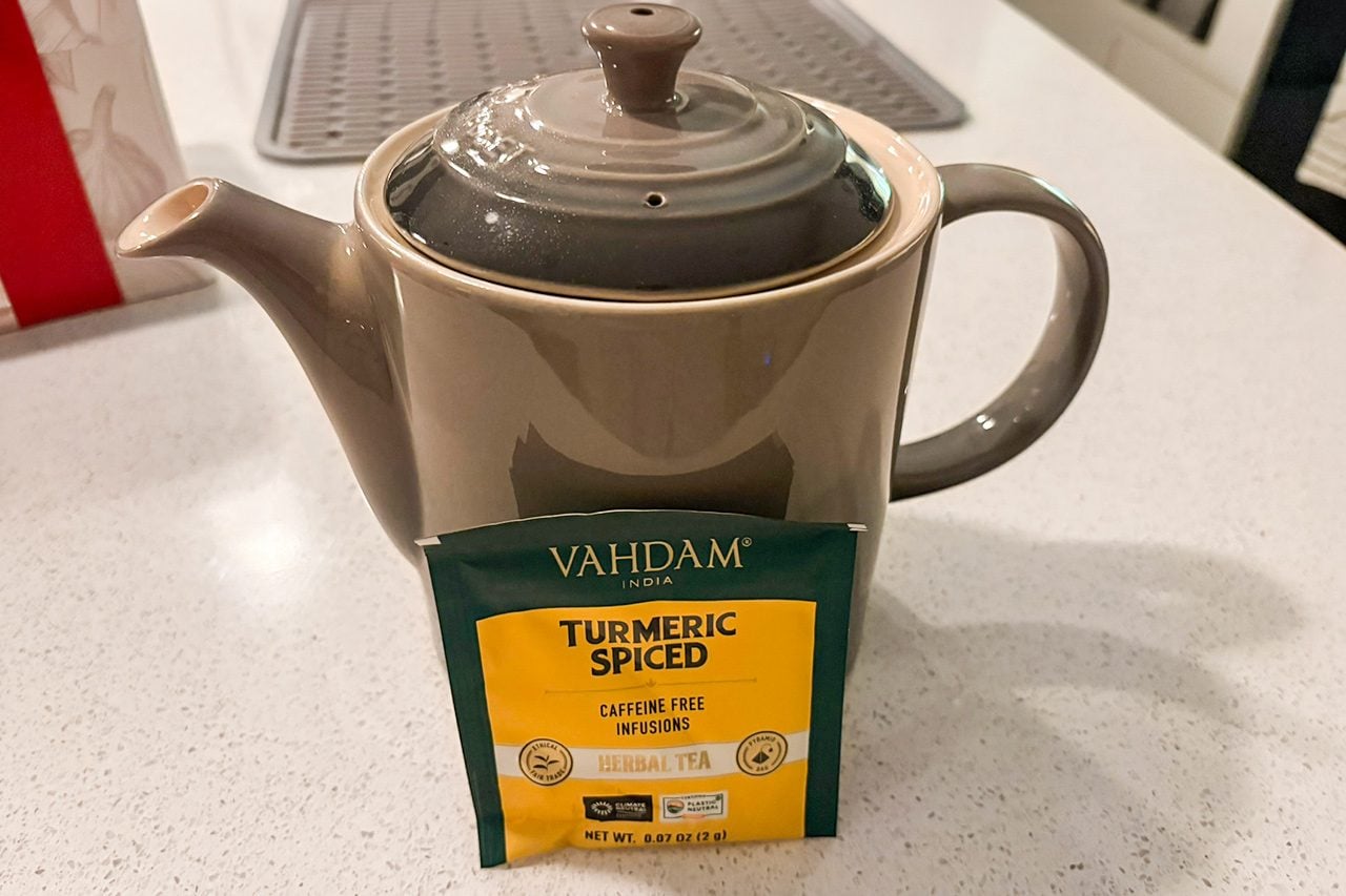 A beige teapot with a lid sits on a white countertop, with a packet of Vahdam Turmeric Spiced tea propped against it.