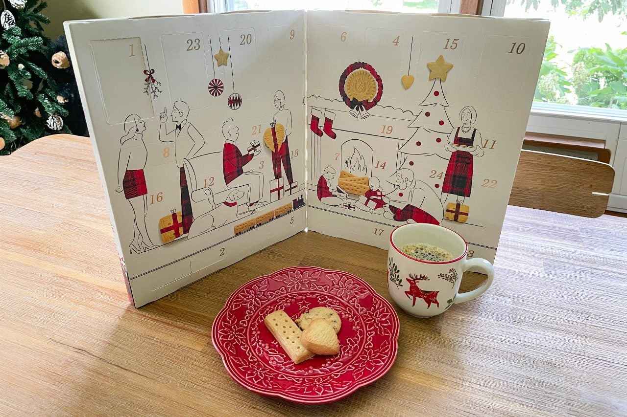 An advent calendar stands open on a table next to a red plate with two cookies and a festive mug. A decorated Christmas tree is visible on the calendar.