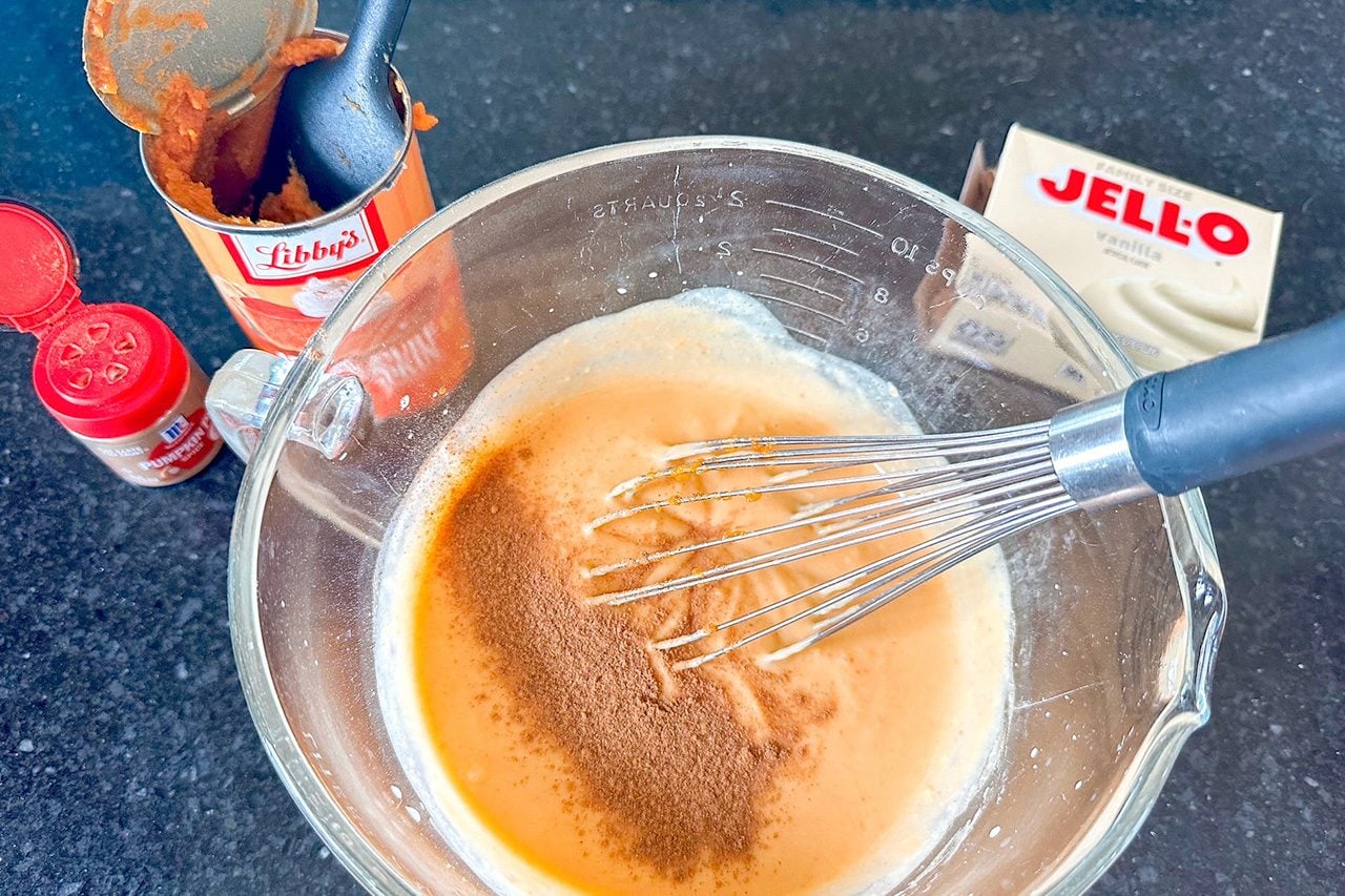 Overhead view of a glass bowl with orange batter, cinnamon, and a whisk, beside Libby’s pumpkin, spices, and Jell-O on a dark counter.