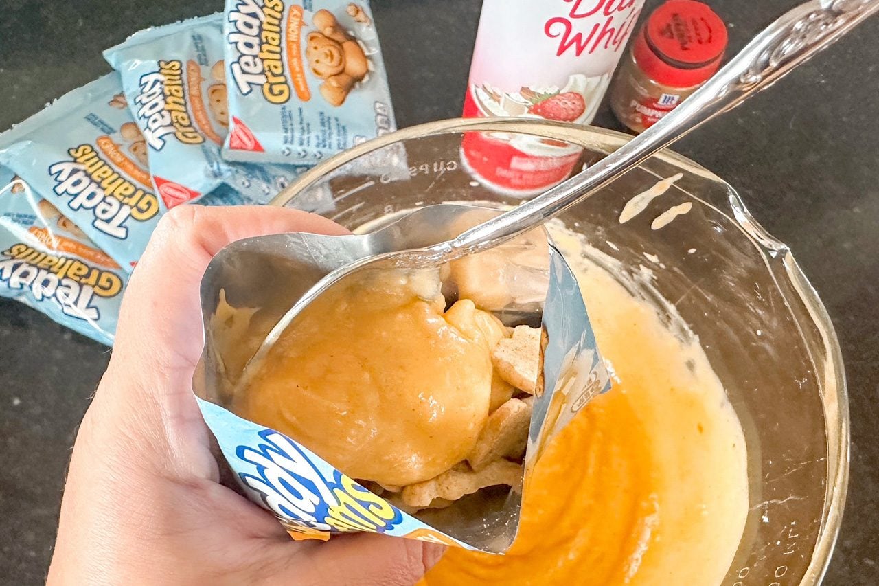 Hand holding an opened snack pouch over a glass bowl of creamy orange dip, with a spoon, croutons, and packaged treats in the background.