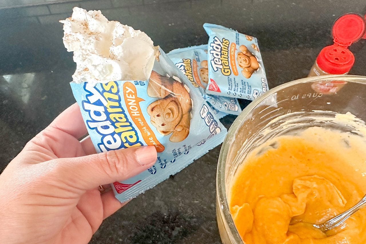 A hand holds opened Betty Crocker cinnamon roll mix packets beside a bowl of orange batter, spoon, and red measuring spoons on a counter.