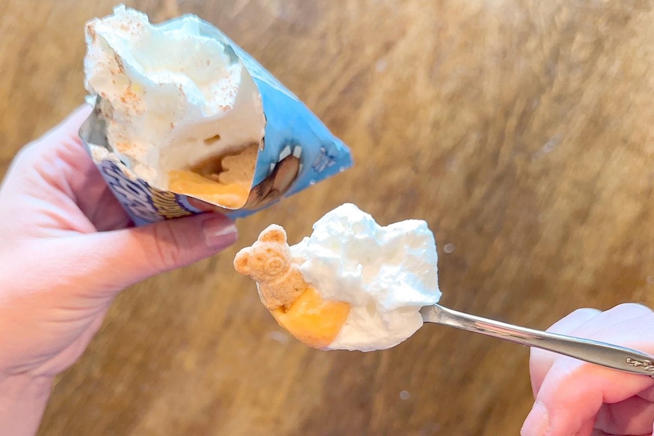 Hands hold a blue-and-yellow dessert wrapper and spoon topped with fluffy white cream and orange filling over a warm wooden table.
