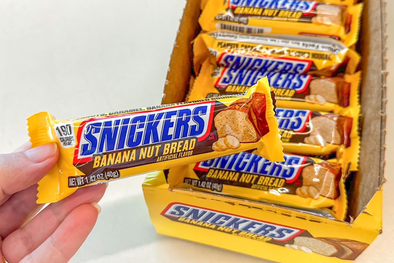 A hand holds a Snickers Banana Nut Bread candy bar. Several more bars are stacked in an open yellow box labeled with the same flavor.