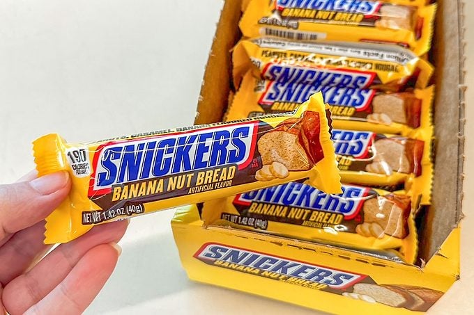 A hand holds a Snickers Banana Nut Bread candy bar. Several more bars are stacked in an open yellow box labeled with the same flavor.