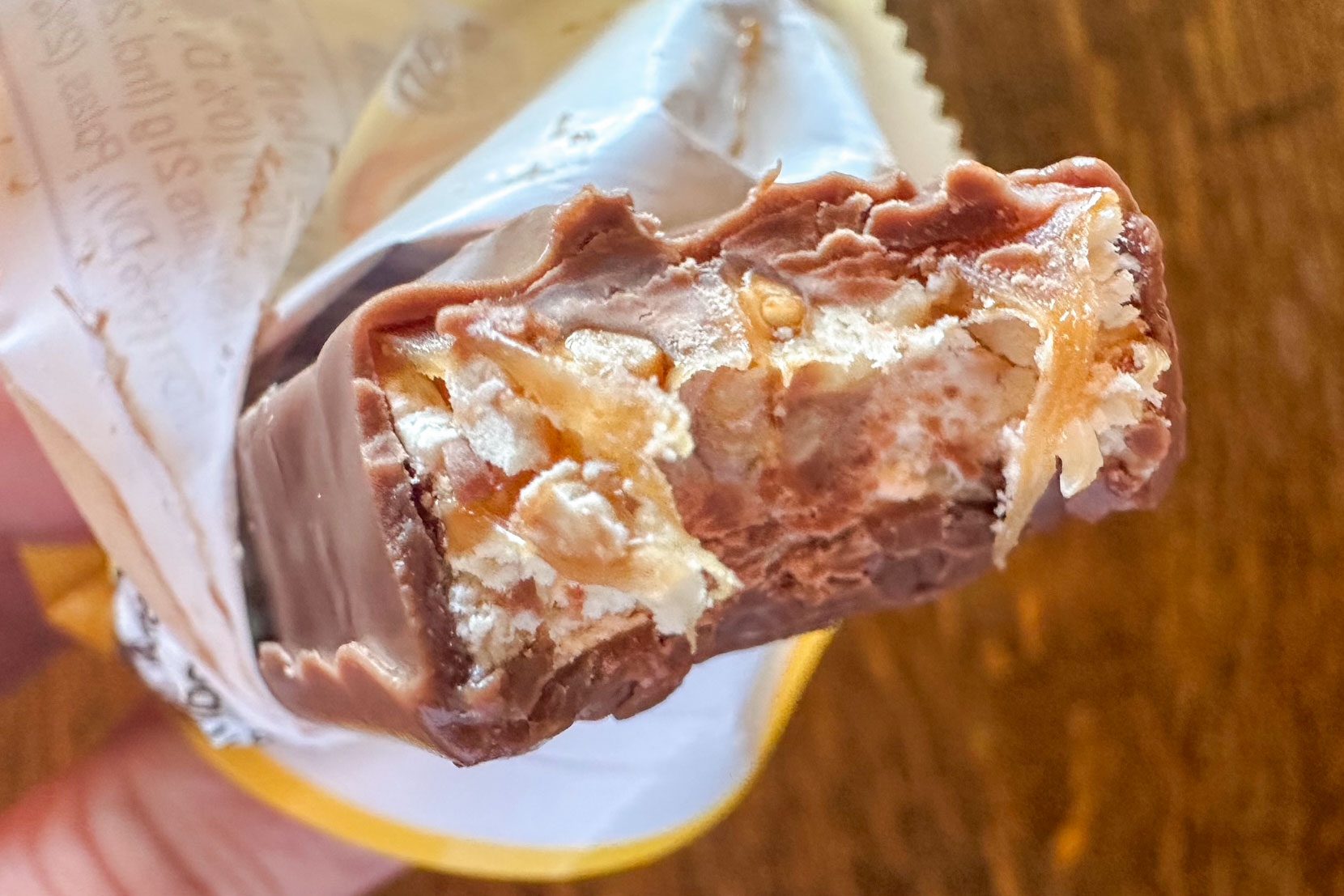 A close-up of a partially eaten chocolate bar, showing layers of caramel, nougat, and peanuts inside, with someone holding the candy in a wrapper.