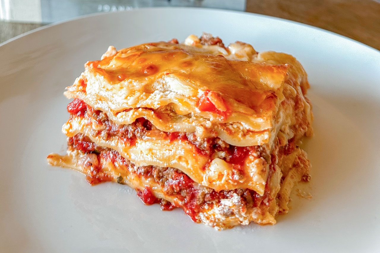 A slice of lasagna sits on a white plate, showing layers of pasta, ground meat, tomato sauce, and melted cheese.