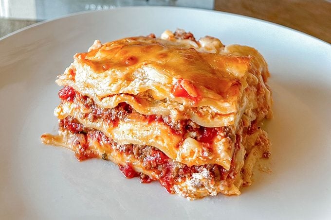 A slice of lasagna sits on a white plate, showing layers of pasta, ground meat, tomato sauce, and melted cheese.