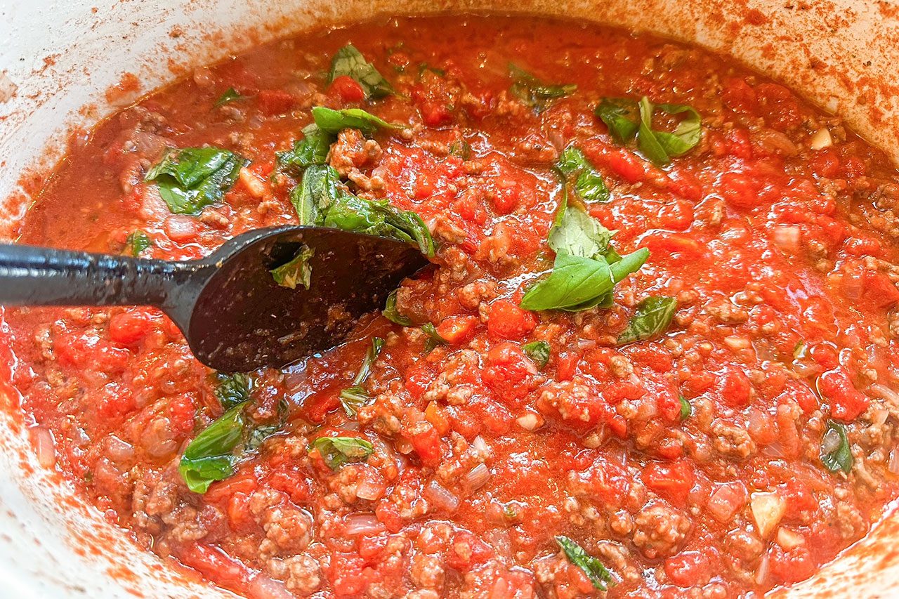 A pot of chunky tomato meat sauce with visible ground beef, tomatoes, herbs, and a black serving spoon stirring the mixture.