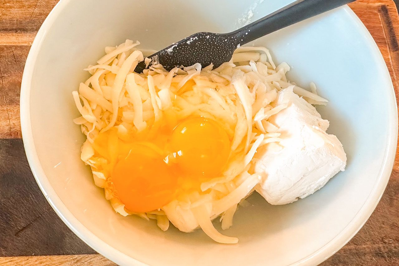 A bowl containing shredded cheese, two raw eggs, and a chunk of cream cheese with a black spatula resting inside.