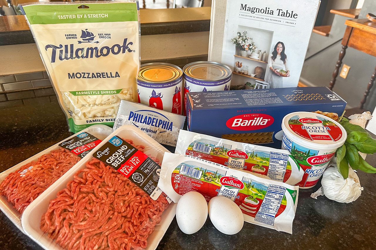 Ingredients for a recipe, including ground beef, mozzarella, ricotta, eggs, canned tomatoes, lasagna noodles, cream cheese, basil, and garlic, are arranged on a kitchen counter 