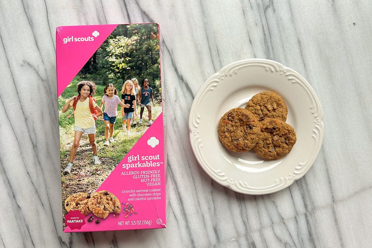 A box of Girl Scout cookies labeled “Sparkle-ables” sits next to a white plate holding three cookies on a marble surface.