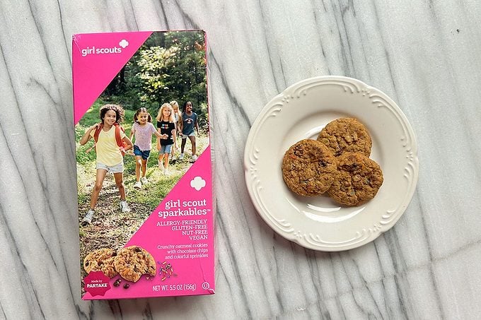 A box of Girl Scout cookies labeled “Sparkle-ables” sits next to a white plate holding three cookies on a marble surface.
