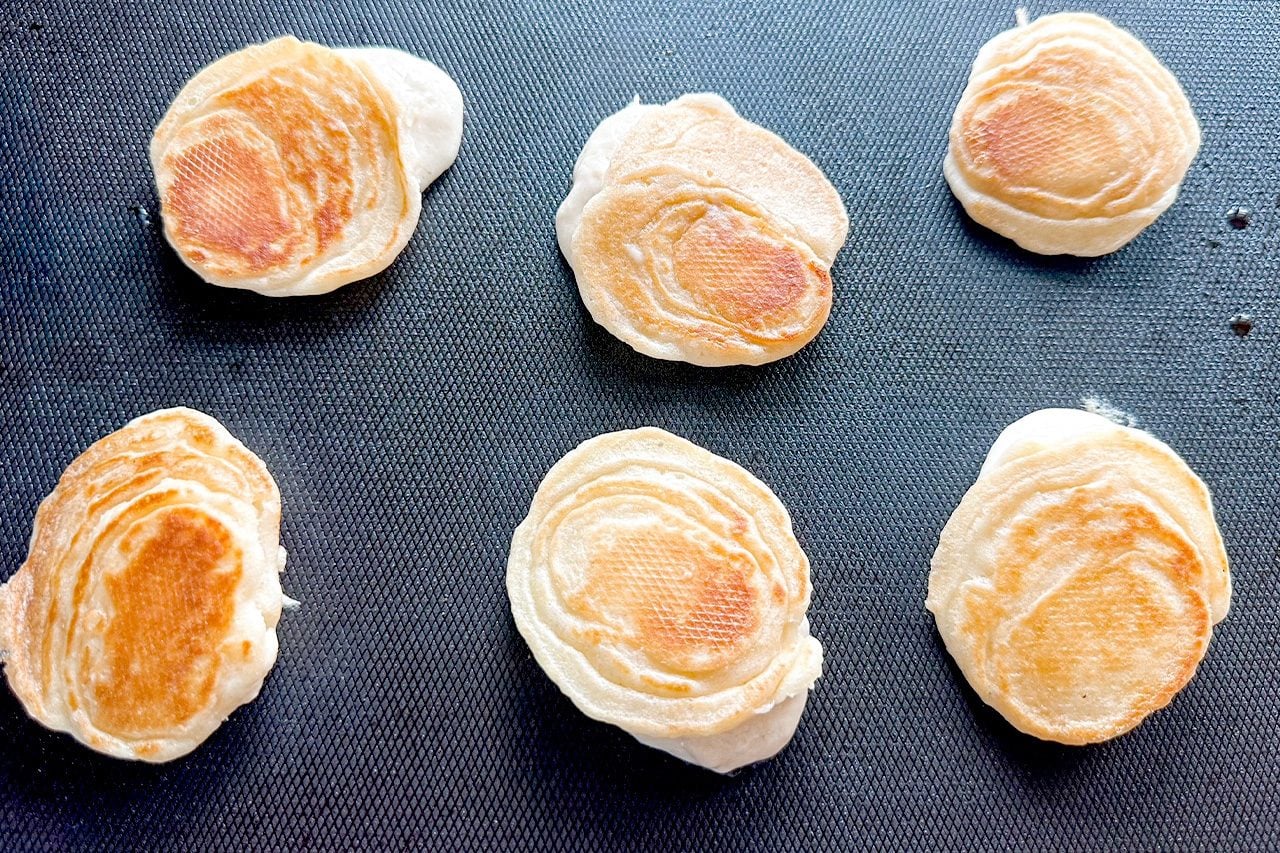 An open hand holds a small, round, golden-brown toasted pastry against dark pavement, creating a casual, everyday scene.