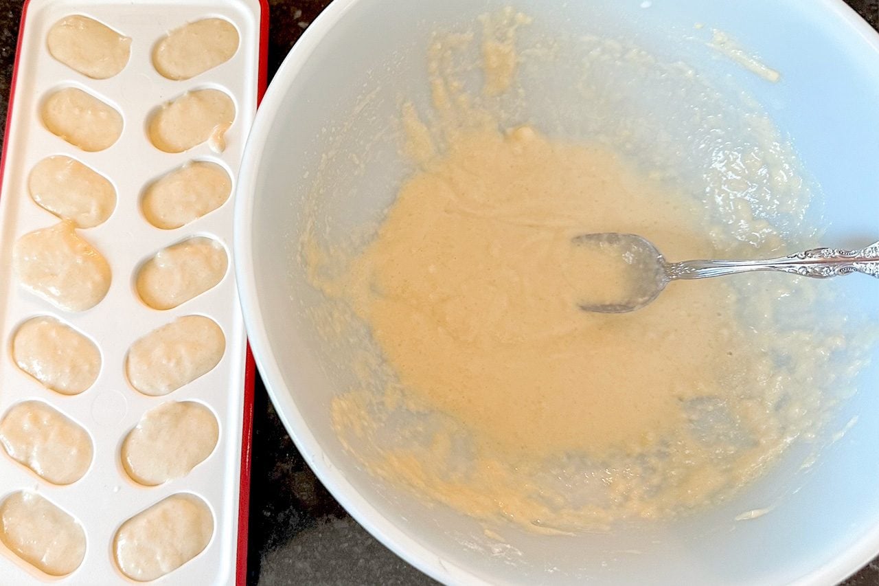 A bowl of pale orange batter with a spoon sits beside a white mold holding rows of small, uncooked dumplings, suggesting homemade cooking.