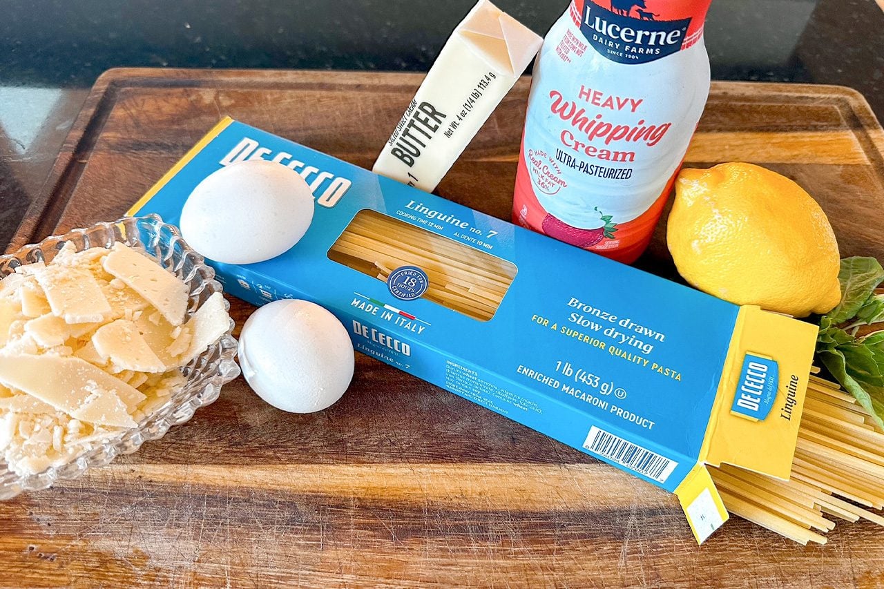 Ingredients for a creamy pasta dish on a wooden board: De Cecco spaghetti, eggs, butter, Parmesan, heavy cream, lemon, and herbs.