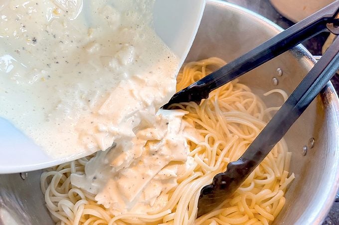 Creamy herb sauce with shredded chicken pours over spaghetti in a metal pan, with black tongs resting on top; a warm, appetizing meal.