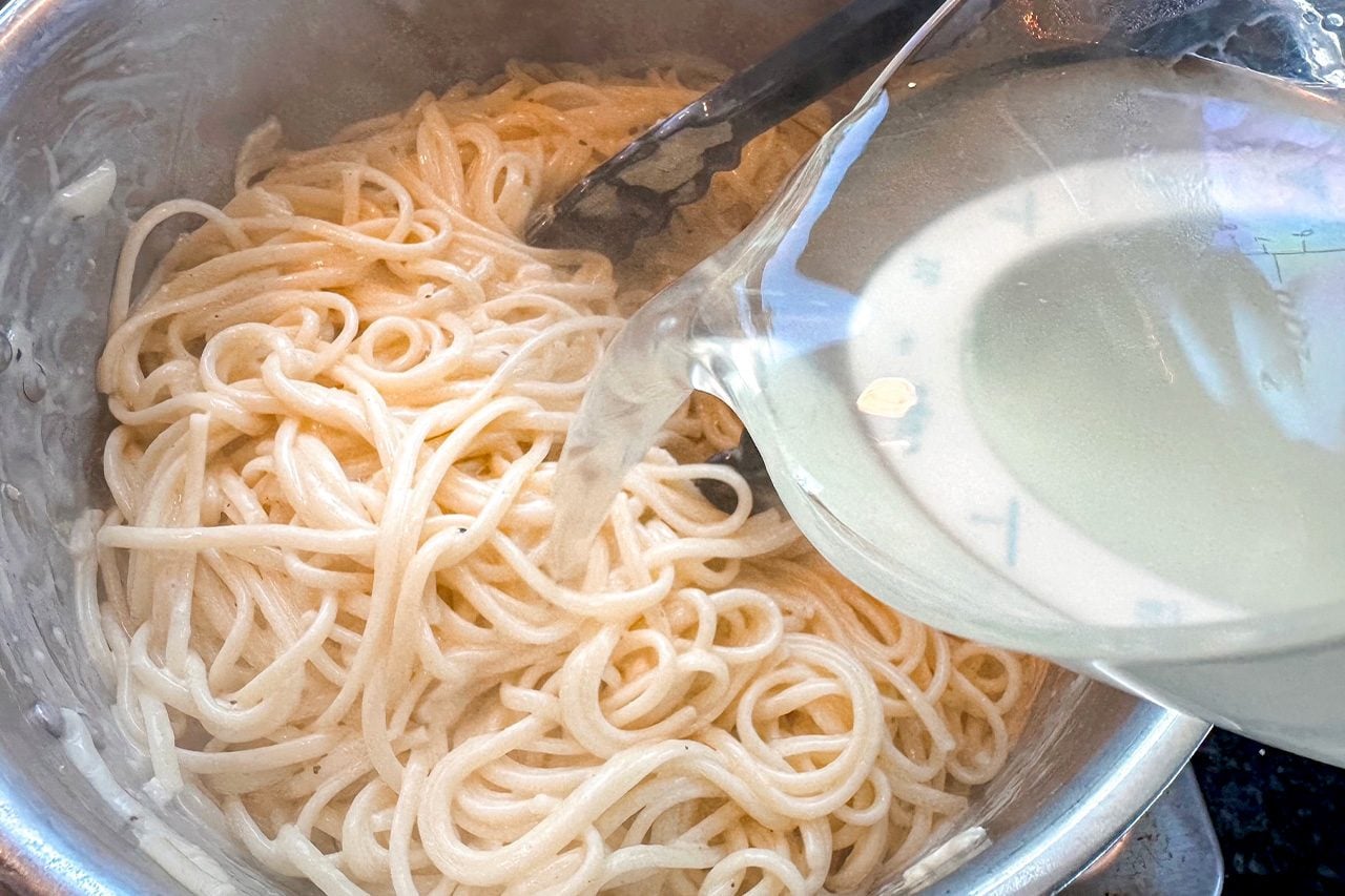 A metal bowl holds plain noodles while tongs rest on top and a glass bowl of pale broth or sauce is tilted over them, suggesting preparation.