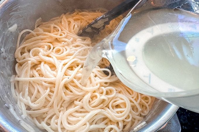 A metal bowl holds plain noodles while tongs rest on top and a glass bowl of pale broth or sauce is tilted over them, suggesting preparation.
