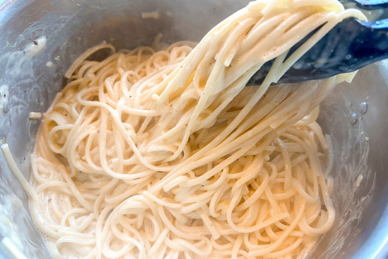 Close-up of creamy spaghetti noodles being lifted with black tongs from a metal bowl, creating an appetizing, warm, homemade cooking scene.